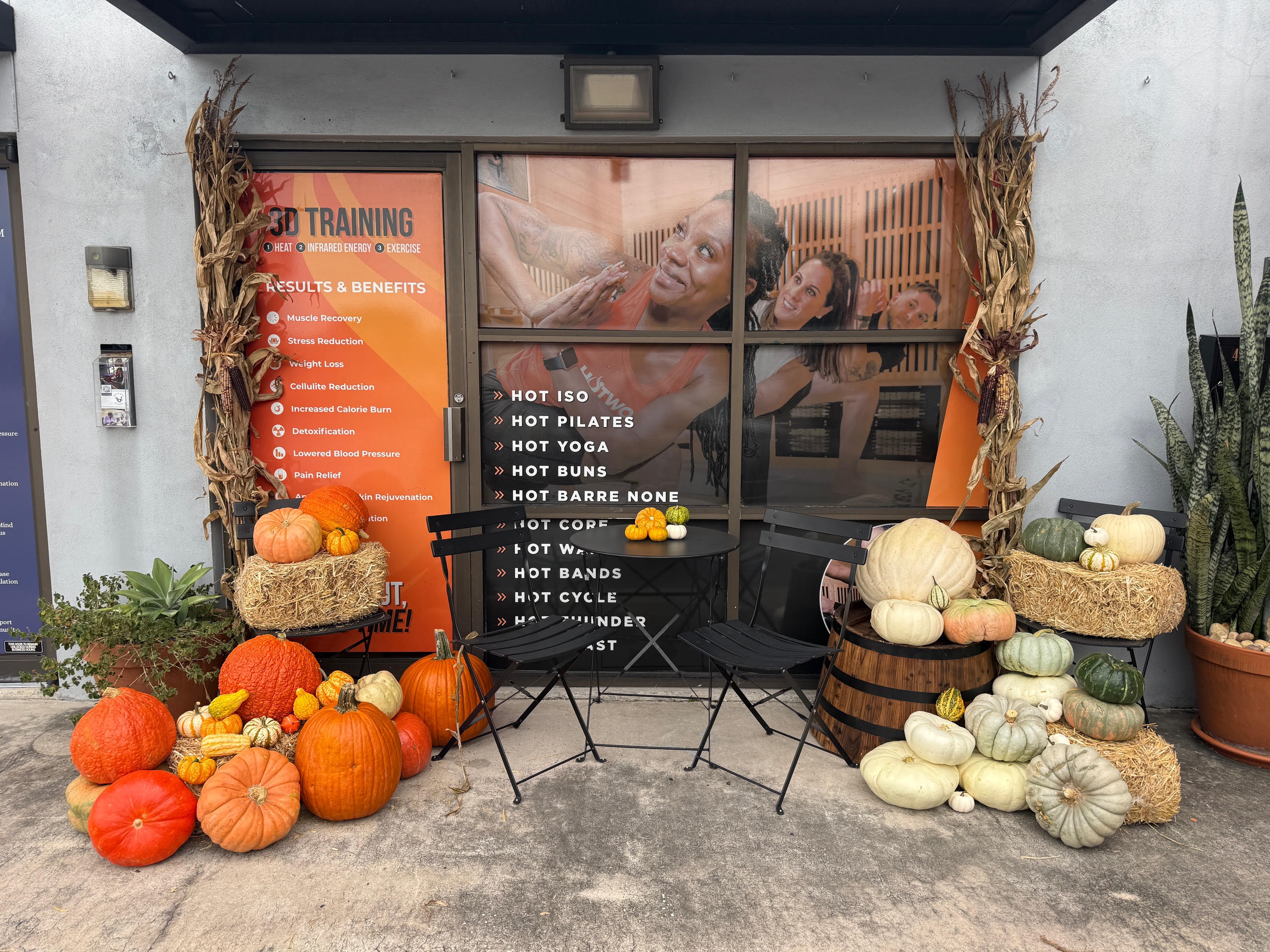 Front-on view of a fall storefront install with symmetrical pumpkin and hay bale arrangements flanking a bistro table